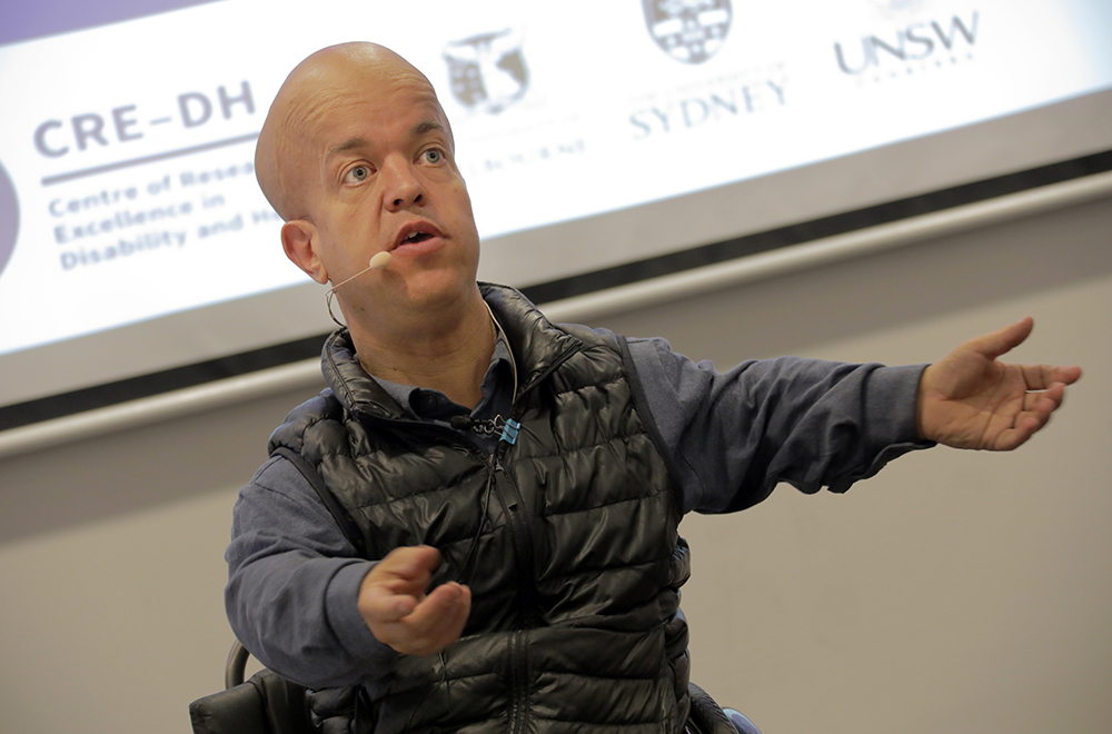 Tom Shakespeare, a small man with a bald head, gestures as he delivers a presentation in front of a screen. He is in a wheelchair and wearing a grey shirt.