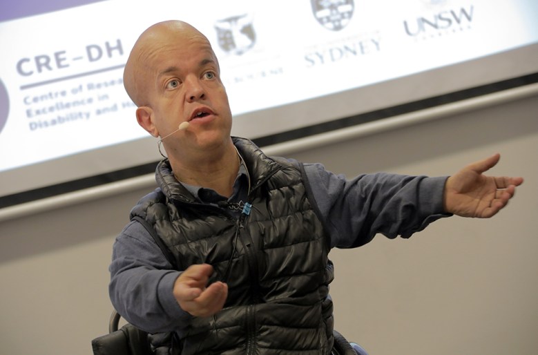 Tom Shakespeare, a small man with a bald head, gestures as he delivers a presentation in front of a screen. He is in a wheelchair and wearing a grey shirt.