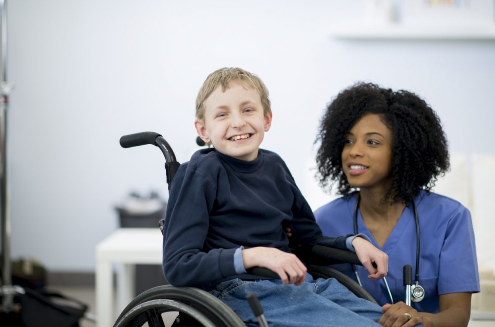 A nurse is with a child in the hospital that has cerebral palsy. The little boy is sitting in a wheelchair. He is smiling and looking at the camera.