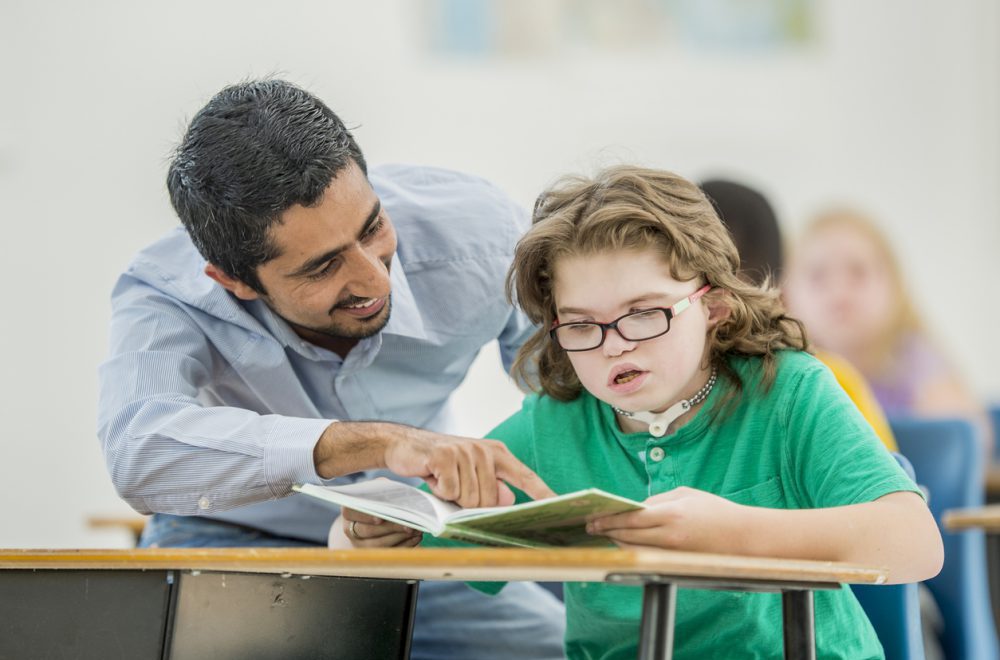 A young girl with Down syndrome reads a book at a school desk. She is wearing a green top. A man is sitting next to her, smiling and pointing at the book.
