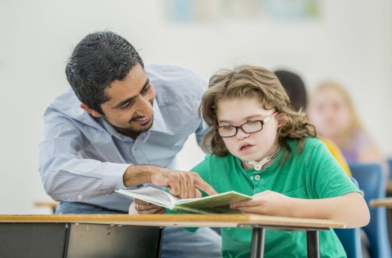 A young girl with Down syndrome reads a book at a school desk. She is wearing a green top. A man is sitting next to her, smiling and pointing at the book.