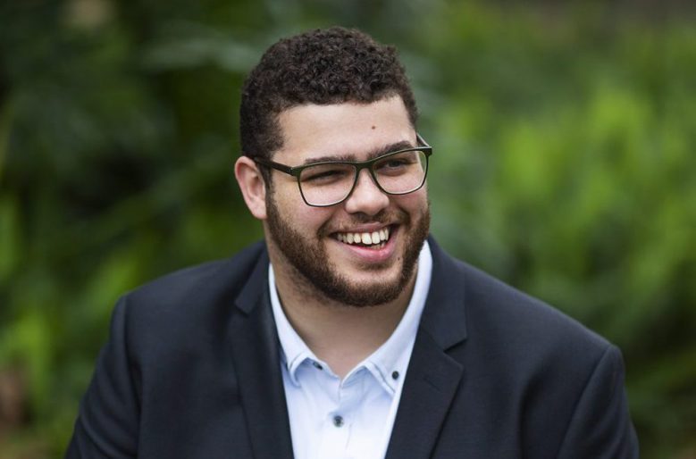 Jordon Steele-John, a senator, a young man with short dark hair and a neat beard, sitting in front of trees. He is smiling warmly. He has glasses.