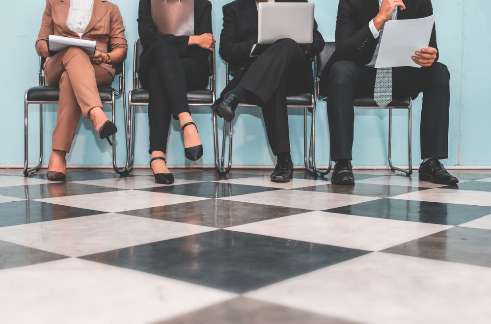 we see a group of people from the knees down, sitting on chairs on a chequered floor.
