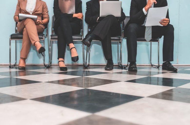 we see a group of people from the knees down, sitting on chairs on a chequered floor.