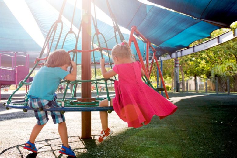 Two children playing at a colourful park. One is a girl in a pink dress, the other is a boy in a blue tshirt. We see them from behind.