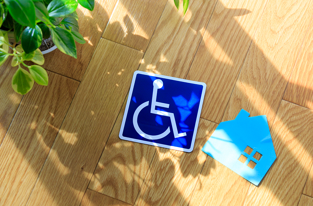 A blue disability sign featuring a white stick figure with a wheelchair on a blue background, next to a blue cutout house, laying on floorboards.