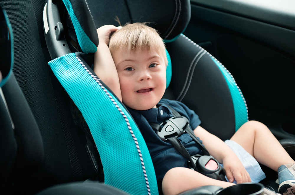 Little boy with Down Syndrome is buckled into car seat. He is smiling and looking the camera. He has blond hair et blue eyes. He wear blue short and sweater. Color and horizontal photo was taken in Quebec Canada.