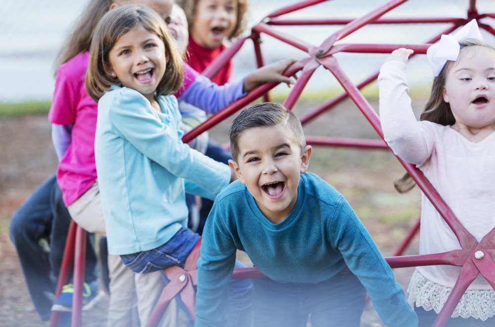 A group of preschool and elementary school children, 5 to 7 years old, on the playground, climbing on monkey bars. The boy in the middle foreground, shouting and looking at the camera is 6. The girl with the white bow in her hair has down syndrome.