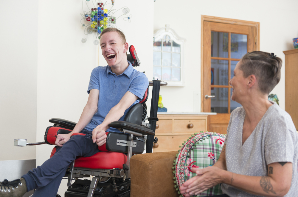 Color image of a real life young physically impaired ALS patient spending time with his mother at home. He is happy.