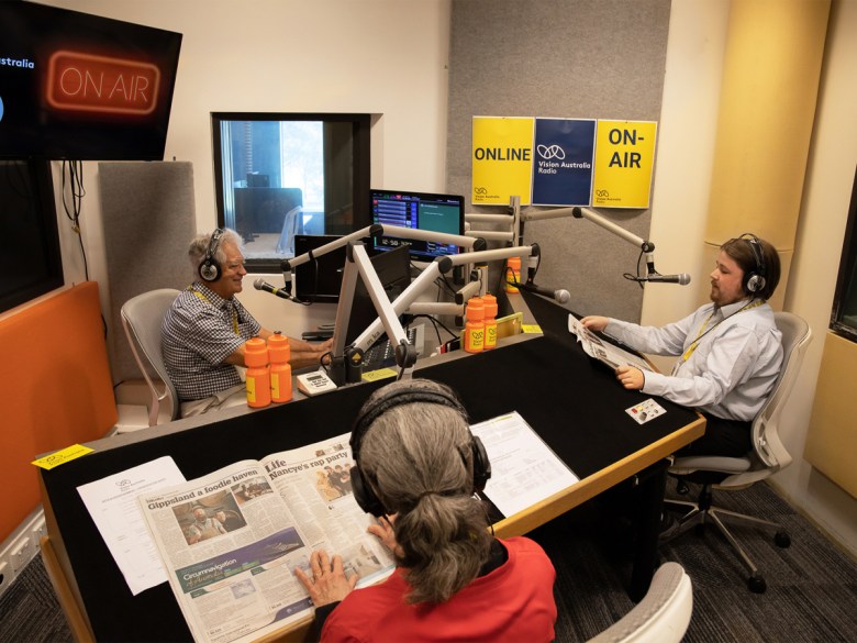 Three people sitting at a desk, wearing headphones in a radio studio. The desks have papers on them.