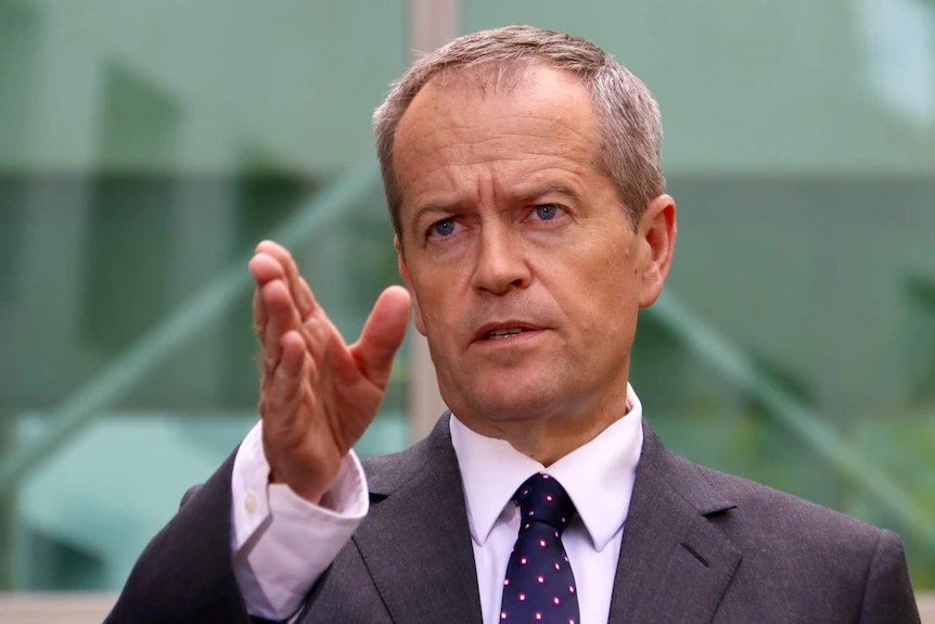 Bill Shorten, a middle aged man with short grey hair, wearing a suit and tie, gestures at the camera.