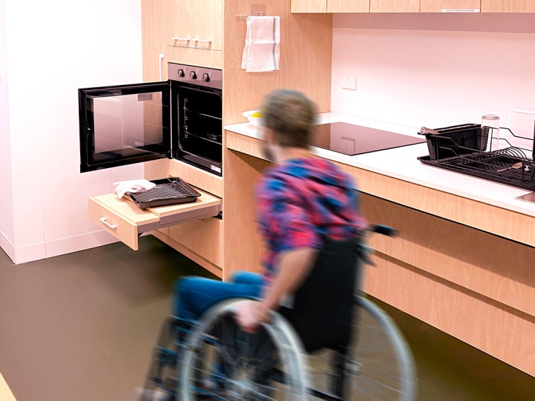 A blurred image of a man using a wheelchair in a specialist kitchen towards a microwave