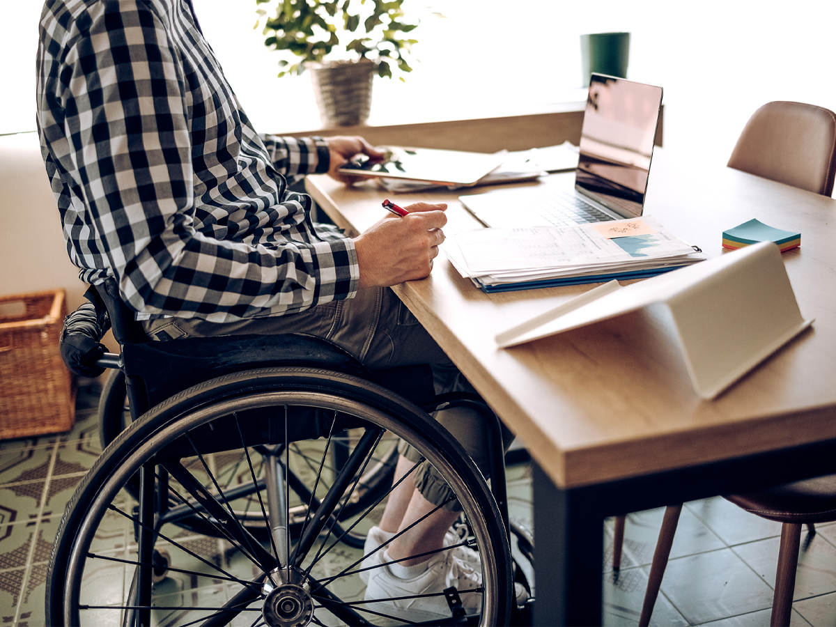 A person sits at a table in a wheelchair. The table is covering in things like books and an iPad.