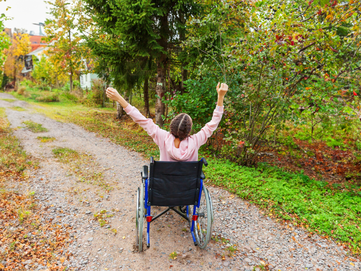 A woman in a wheelchair raises her arms in joy. We see her from the back. She is on a path in a garden.