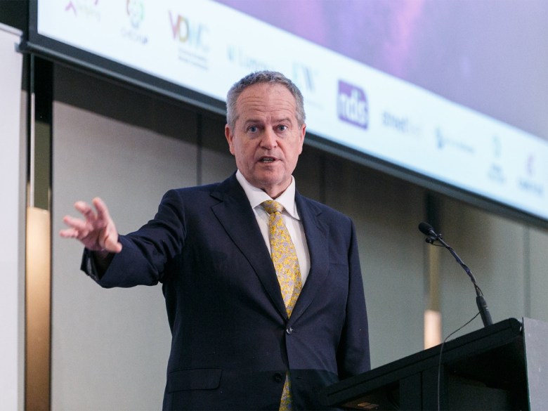 NDIS Minister Bill Shorten, a white man with short receding hair, wears a black suit and yellow tie and is speaking at a lectern.