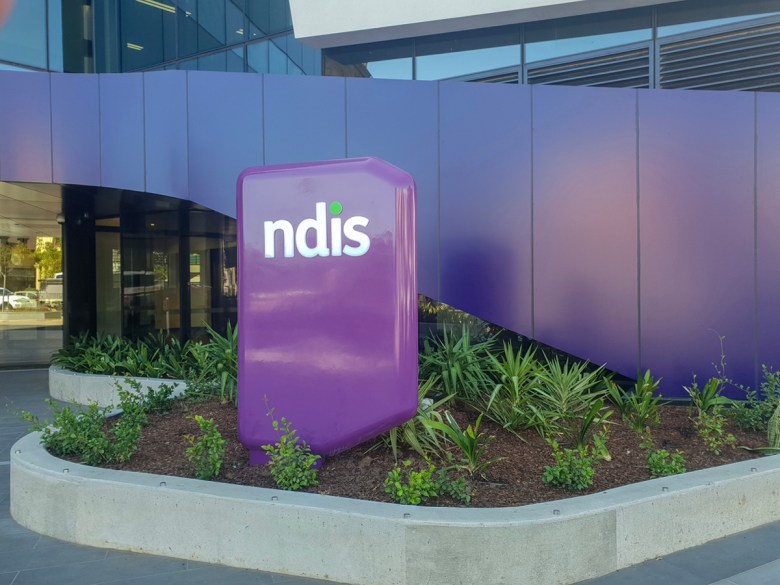 A purple sign with the word NDIS in white written on it. It sits in a garden bed in front of a purple sloping wall.