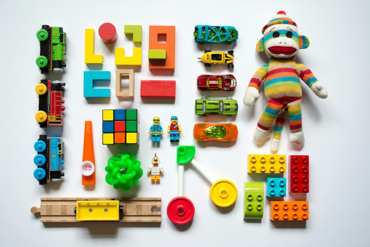 A series of colourful children's toys lined up on a white surface. There are lego blocks and trains and a doll among them.