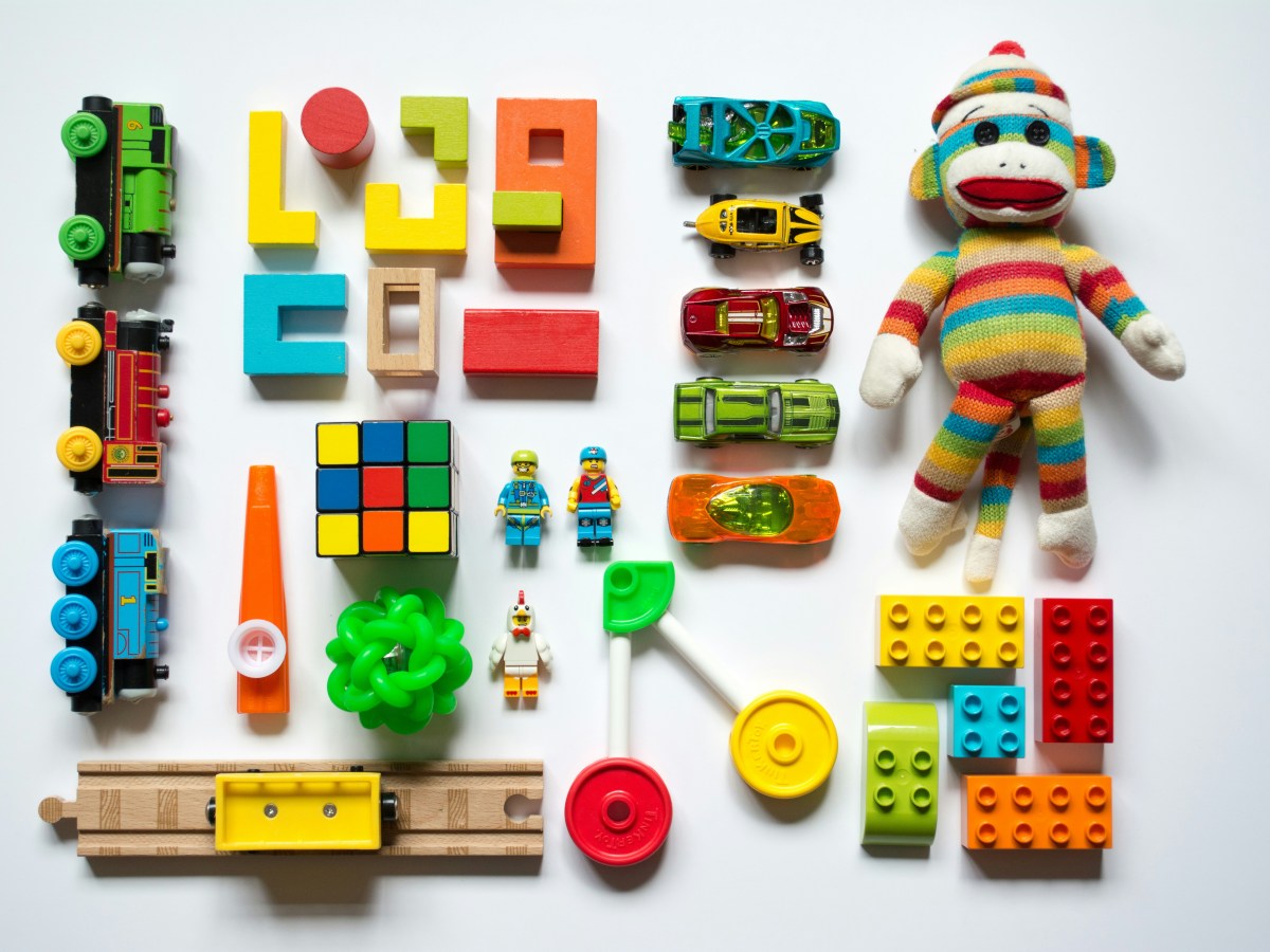 A series of colourful children's toys lined up on a white surface. There are lego blocks and trains and a doll among them.