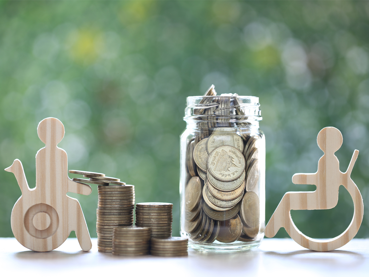 Wooden ornaments of people in wheelchairs next to a jar and stacks of coins, sitting on a table with a green background.