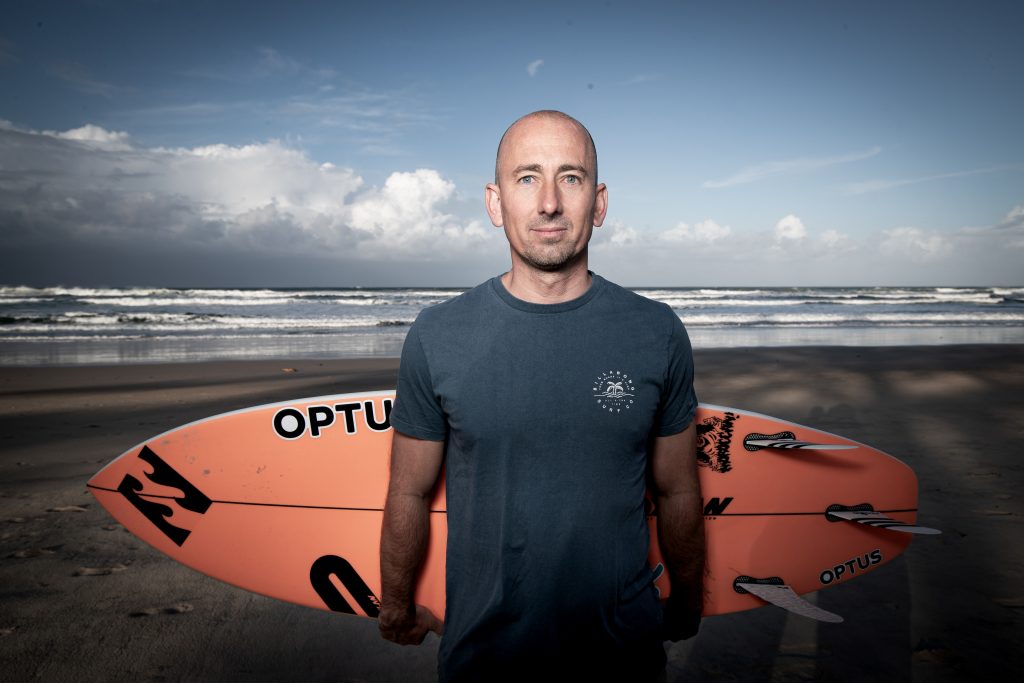 Matt Formston, a surfer with disability, stands on a beach with a surfboard behind him. He is bald and wearing a blue tshirt.