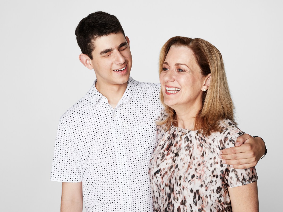 A teenage boy with dark hair wearing a pale shirt, with his arm around his mum, a middle aged woman with blonder hair and a patterned shirt.