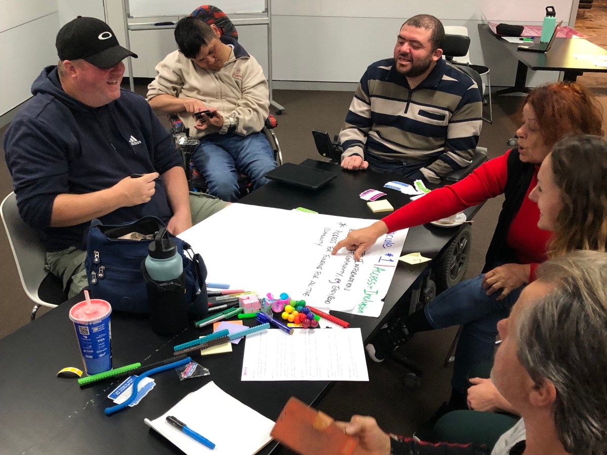 A group of people, some with a physical disability, sit in a room around a table with documents on it. They are having a discussion.