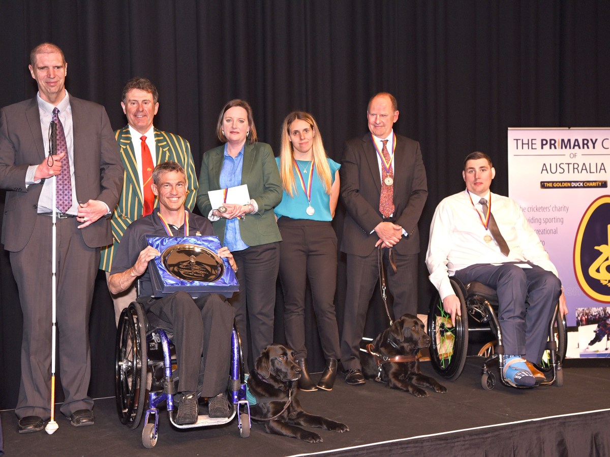A group of people at a previous Primary Club dinner. They are on a stage and several people have a visible disability, including two people in a wheelchair and one who uses a white cane.