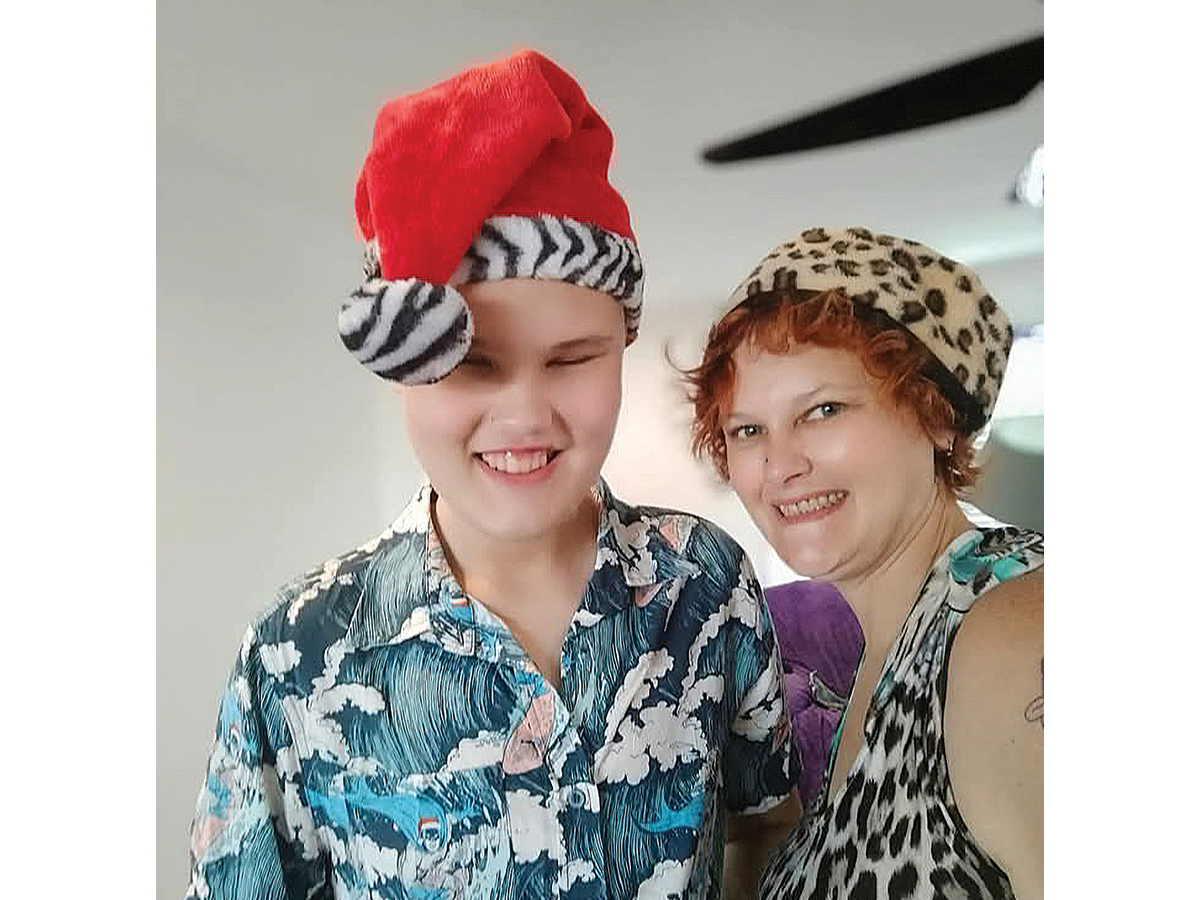 A young boy in a floppy red hat and blue patterned shirt smiles with an older lady with bright red hair.