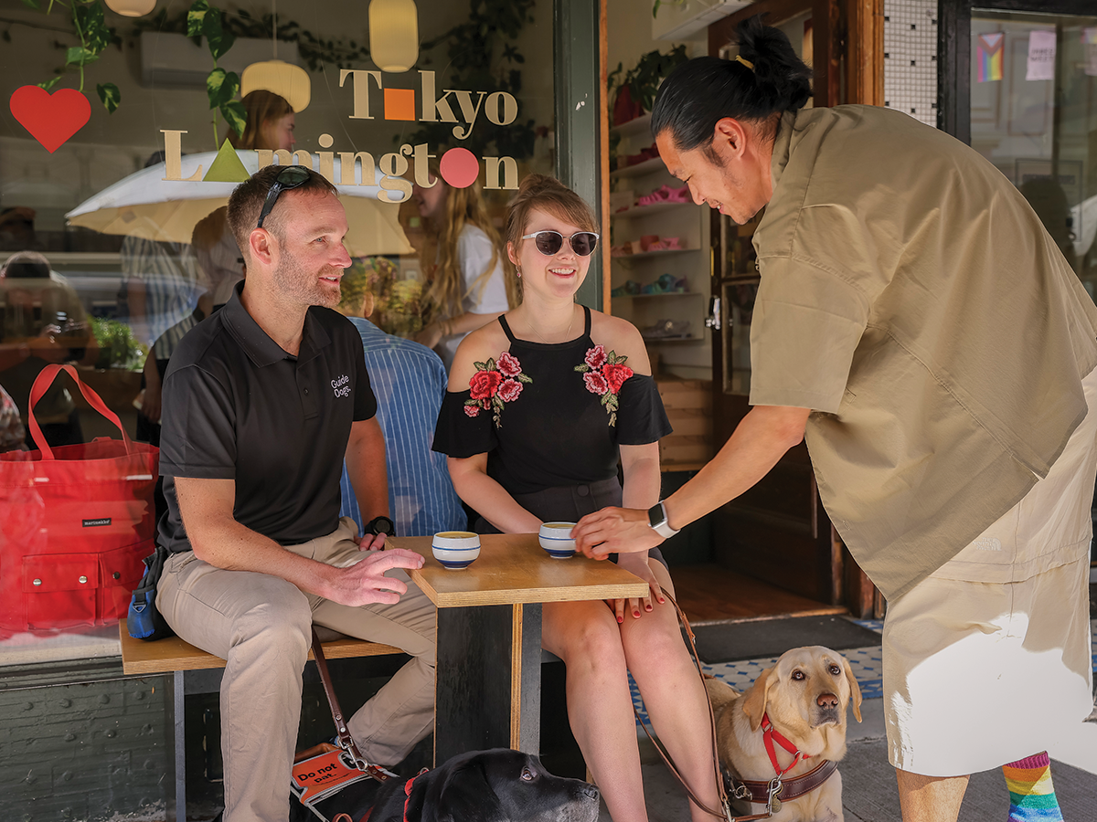 Two people, a man and a woman, are sitting outside a cafe. A waiter bends in front of their table to give something to them.
