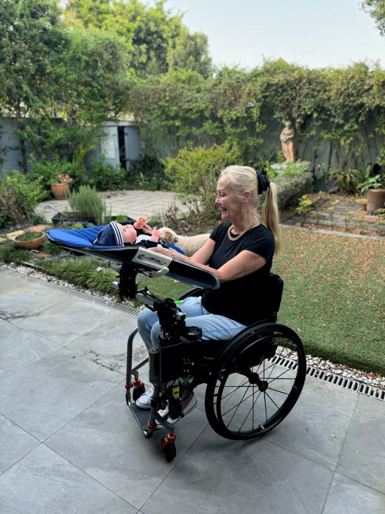 A lady in a wheelchair uses a Care Station, a platform attached to her chair for her baby. She is in a garden setting.