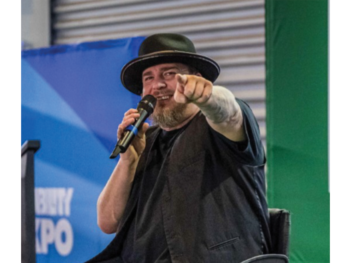 Mick on Wheels, a man in a wheelchair holding a microphone wearing a black hat. He is pointing to the camera.