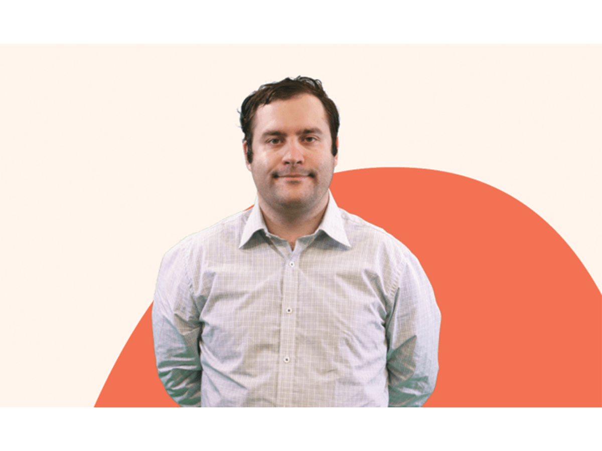 Michael Theo, a young man in a light coloured business shirt with dark short curly hair, stares at the camera. There is an orange background.