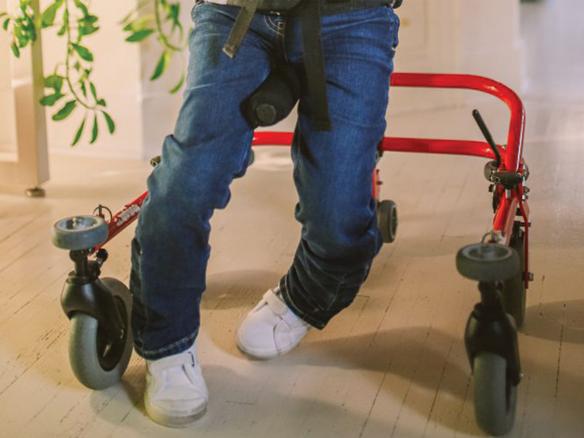 We see the bowed legs of a child with cerebral palsy, in a walking frame. They are wearing blue jeans and white shoes.