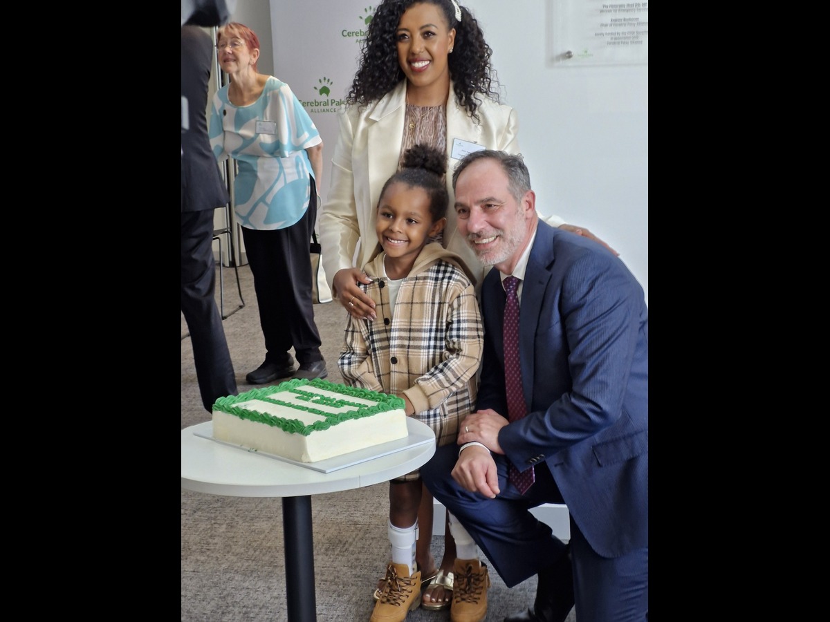 A young girl stands at a table with an older man next to her. They are both smiling. A lady stands behind the girl and has a hand on her shoulder. It is a happy photo.