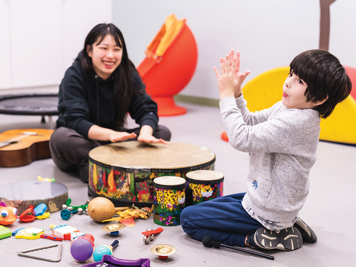 An Asian woman sits on the ground in the background amongst music toys, smiling at a young boy with dark hair who is clapping.