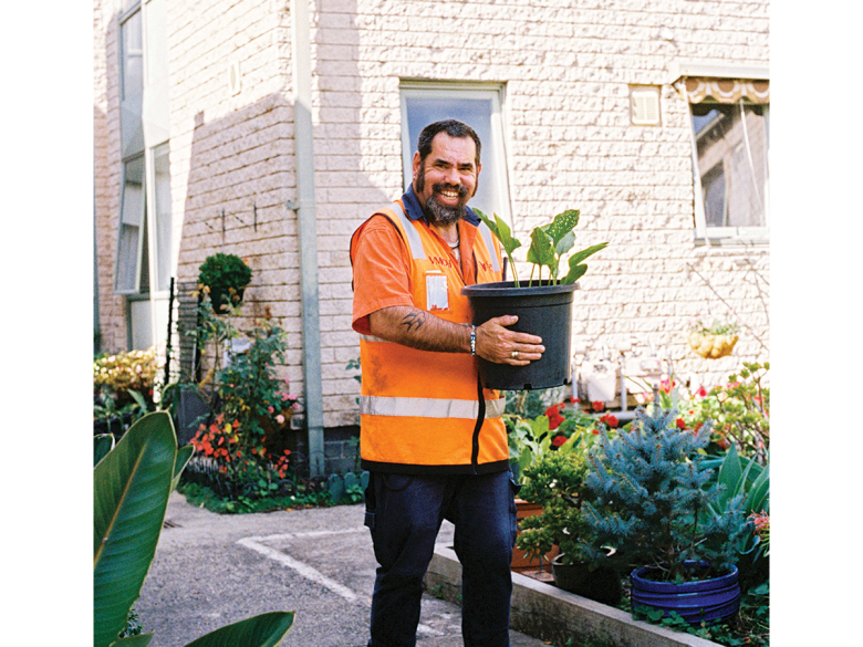 A man with a thick brown beard in an orange high vis vest carrying a plant in a black pot through a courtyard garden. He is smiling and it is sunny