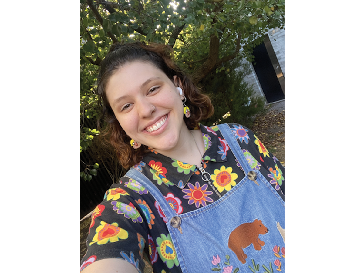 A young woman in a colourful floral shirt and blue overalls smiles in a selfie. She has brown shoulder length hair.