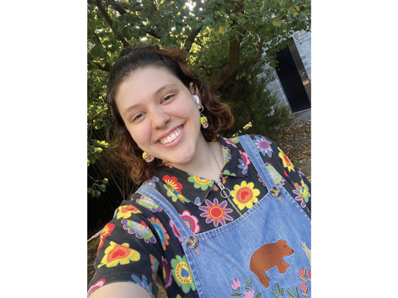 A young woman in a colourful floral shirt and blue overalls smiles in a selfie. She has brown shoulder length hair.