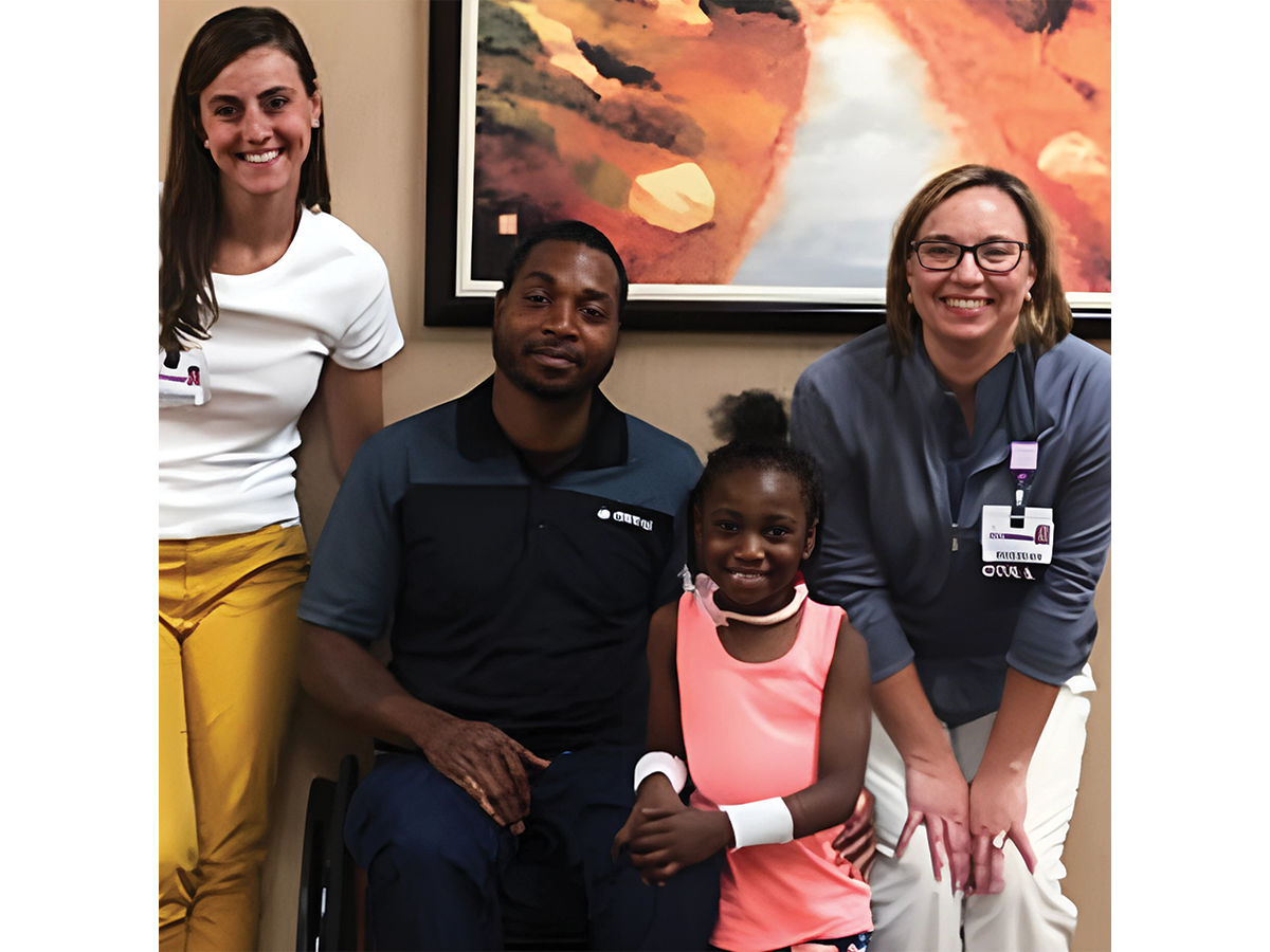 A black man who users a wheelchair sits in the centre, dressed in dark clothes. On his right is a young girl with long dark hair in a white tshirt, and on the other side is a little girl in pink and an older lady with an official badge on and short, flat brown hair.