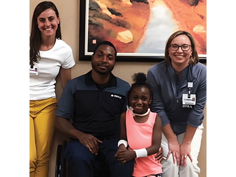 A black man who users a wheelchair sits in the centre, dressed in dark clothes. On his right is a young girl with long dark hair in a white tshirt, and on the other side is a little girl in pink and an older lady with an official badge on and short, flat brown hair.