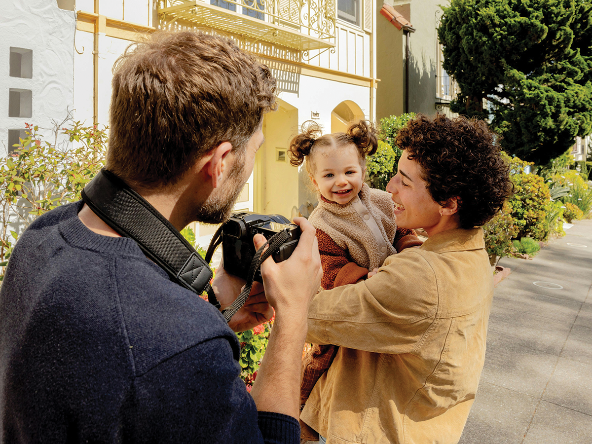 A young boy in a tan shirt holds a little girl in pigtails who is smiling, while a photographer points a camera at them. We are looking over the shoulder of the photographer. They are all outside.