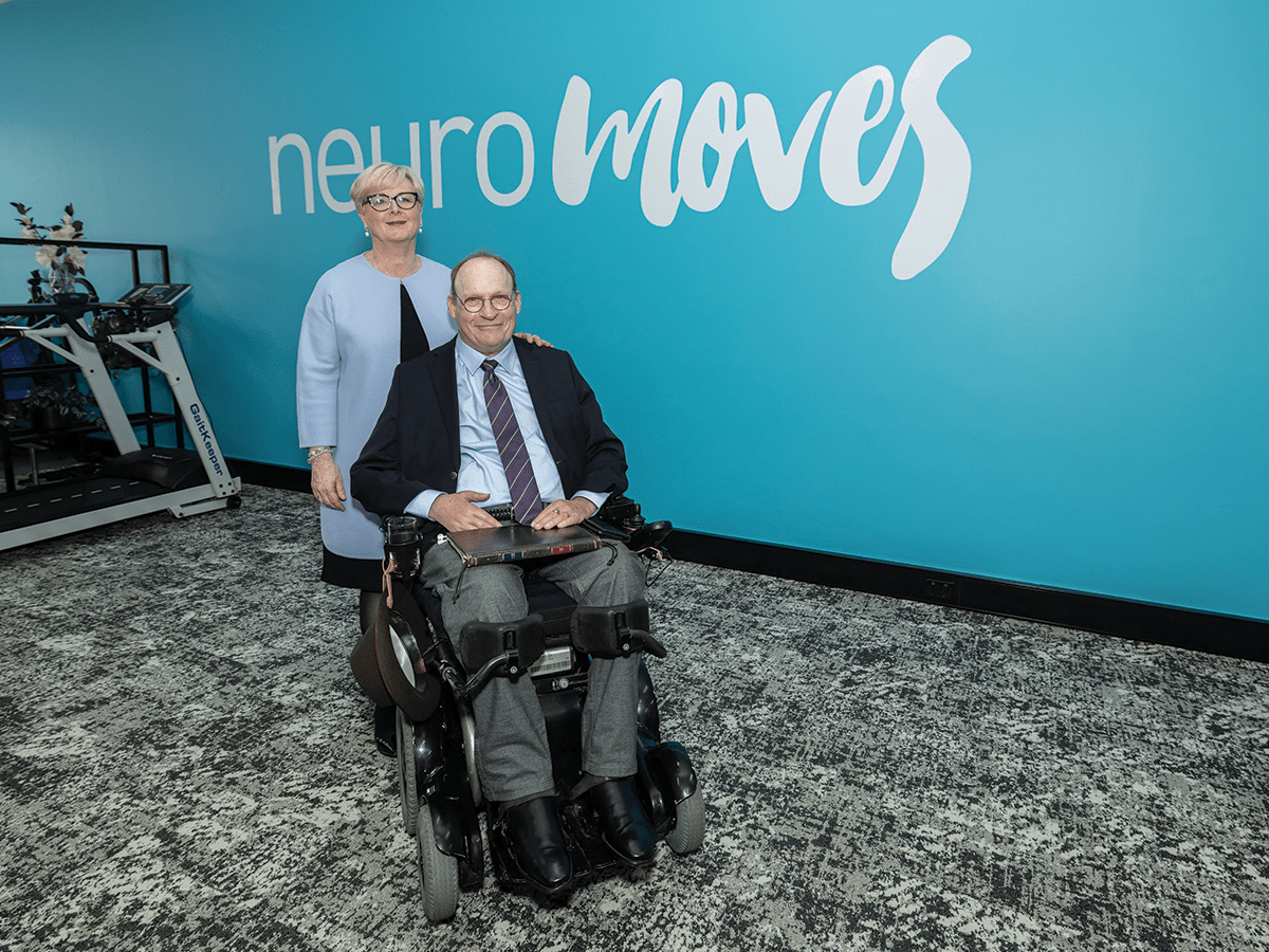 A lady in a blue blazer with short white hair stands behind a man in a black blazer in a wheelchair, in front of a blue wall that says Neuromoves