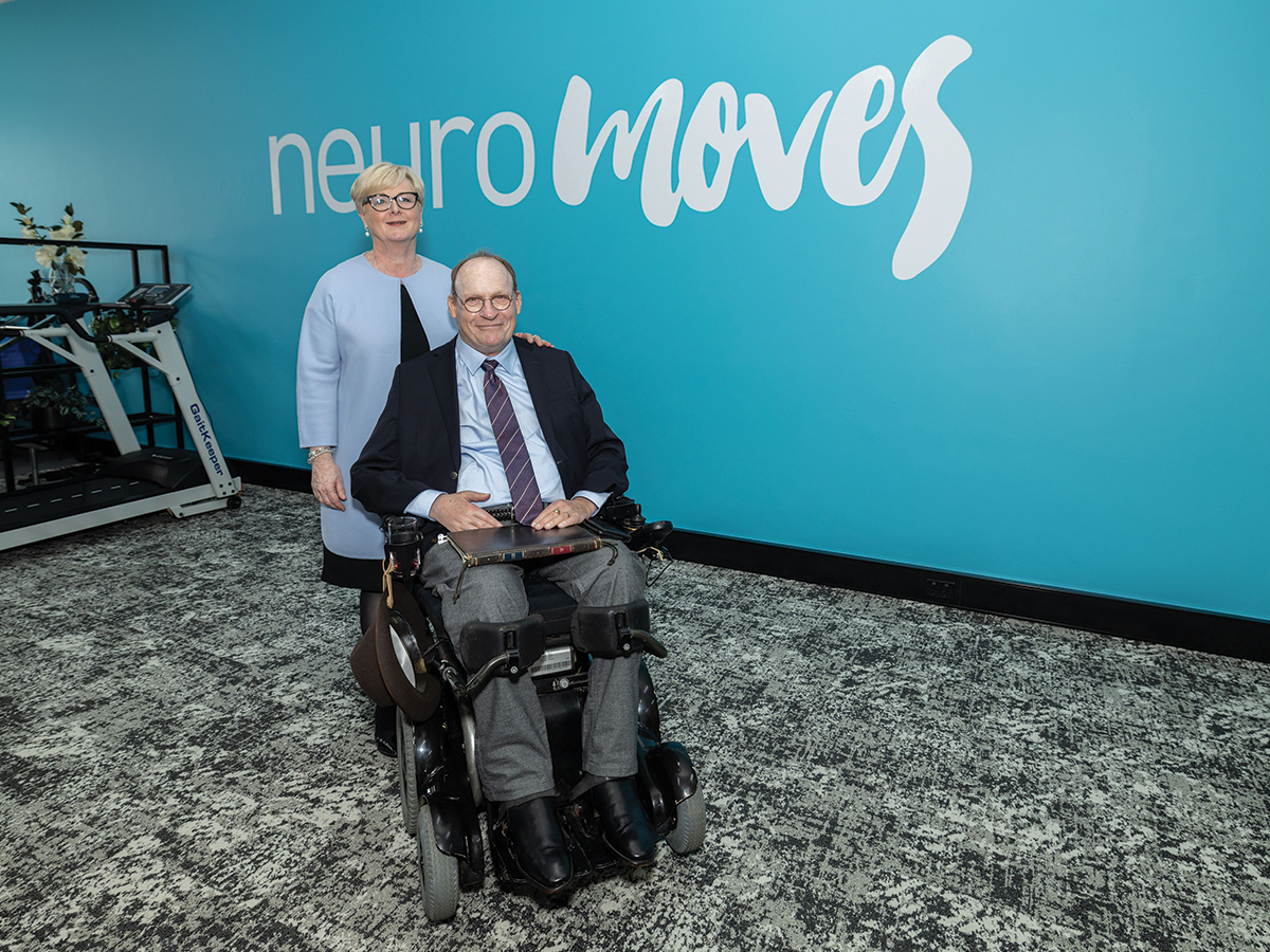 A lady in a blue blazer with short white hair stands behind a man in a black blazer in a wheelchair, in front of a blue wall that says Neuromoves