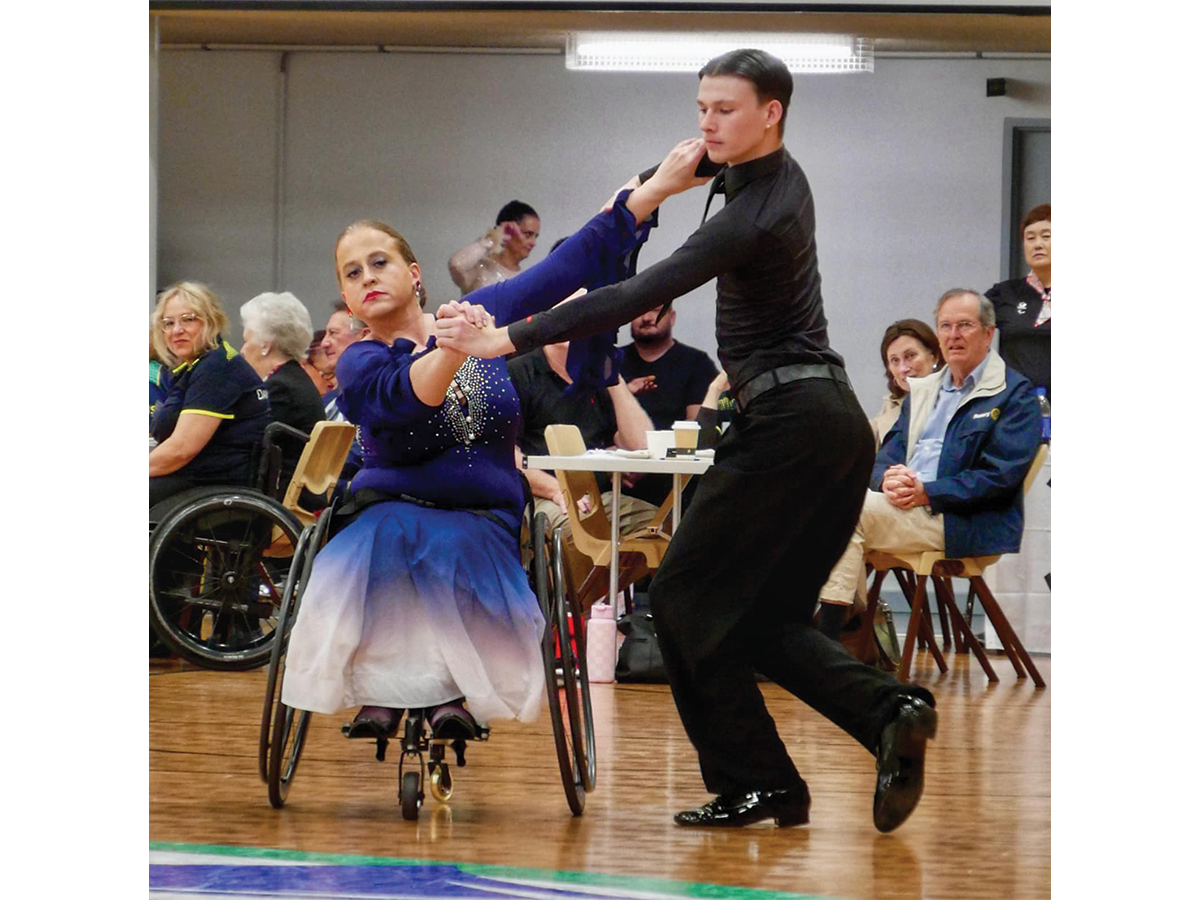 Two para dancers, one a young woman in a sparkly navy dress in a wheelchair, and a young man dressed in black who is standing up, dance together while a crowd watches.