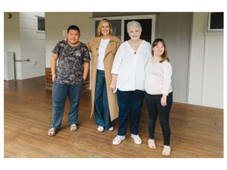 Four people stand in a room, their arms around each other. One is a teenage boy, one is a tall blonde lady, one is an elderly white haired lady, and one is a young brown haired girl. They are smiling.
