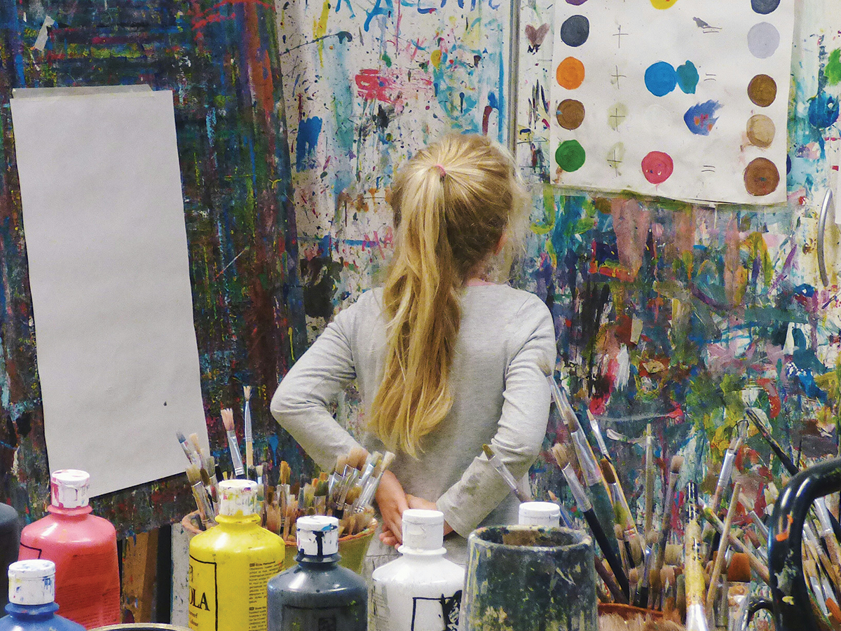 A blonde lady, seen from behind, in a paint studio, looking at colourful paint on a wall.