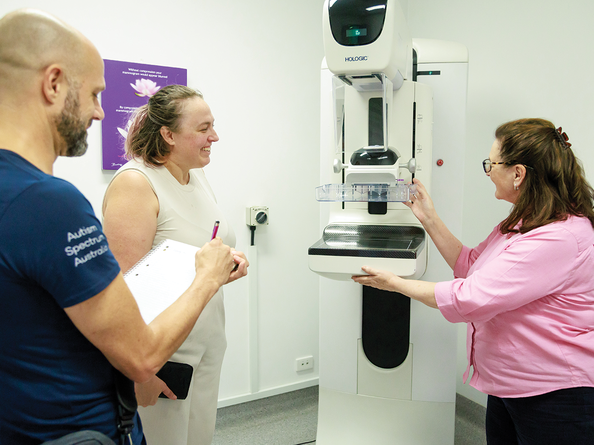 Three people surround a mammogram machine. One is a blonde woman in a white singlet. One is a bald man in a blue medical shirt. The other is a brunette woman in a pink shirt.