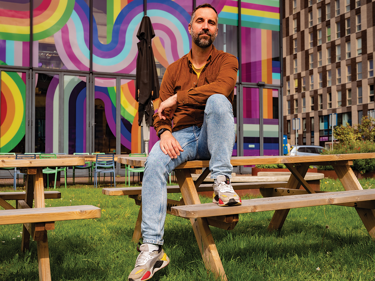 A male in an orange shirt and blue jeans sits on a picnic bench against a vibrant colourful background,