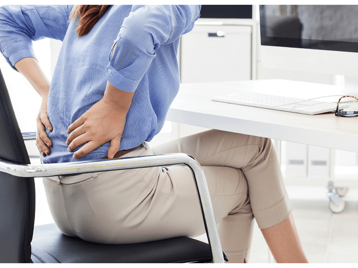 A woman in a blue top and beige pants sits on a chair at a desk, holding her lower back in a sign of chronic pain.