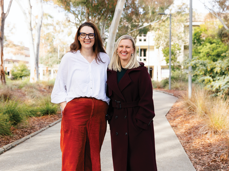 Two women smile brightly at the camera, in a garden. The one on the left is in a white shirt and red pants and has brown hair, the one on the right has blonde shoulder length hair and is wearing all black clothes.